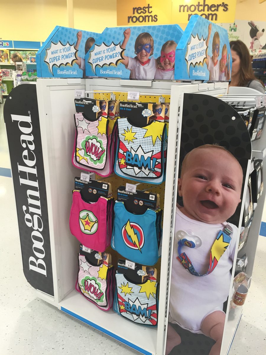 A branded BooginHead Super Power endcap at Babies “R” Us, stocked with bib-and-cape sets and PaciGrip pacifier clips.